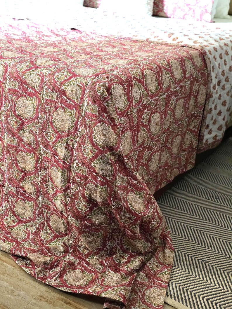 May include: A bedspread with a floral pattern in shades of red, beige, and green. The design features large, stylized flowers and intertwining vines. The bedspread is draped over a bed, with a white sheet and patterned pillows visible.