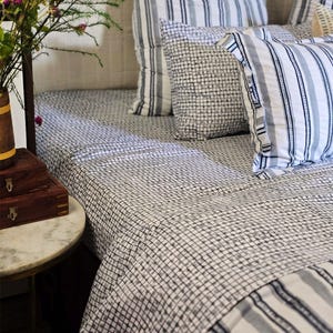 Peut inclure: Un lit avec une couverture et des oreillers à motifs. La couverture a un motif quadrillé, tandis que les oreillers présentent des rayures et un motif quadrillé. Une petite table de chevet avec un plateau en marbre et une boîte en bois sont au premier plan.