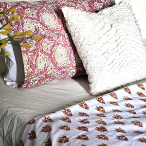 May include: A bed with a white sheet, patterned pillows, and a patterned blanket. The pillows have a pink and white floral design, and a white fluffy pillow. The blanket has a white background with a repeating leaf pattern.