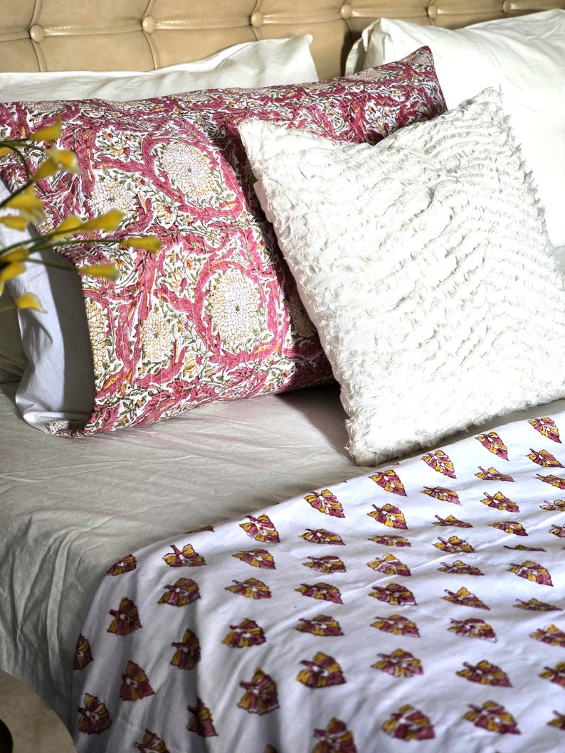 May include: A bed with a white sheet, patterned pillows, and a patterned blanket. The pillows have a pink and white floral design, and a white fluffy pillow. The blanket has a white background with a repeating leaf pattern.
