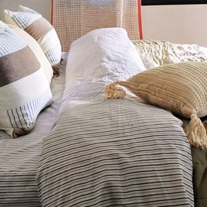 May include: A bed dressed with striped bedding and multiple pillows. The pillows feature diverse patterns, including stripes and colour blocks in neutral tones. A textured pillow with a tassel is also visible. A patterned curtain hangs in the background.
