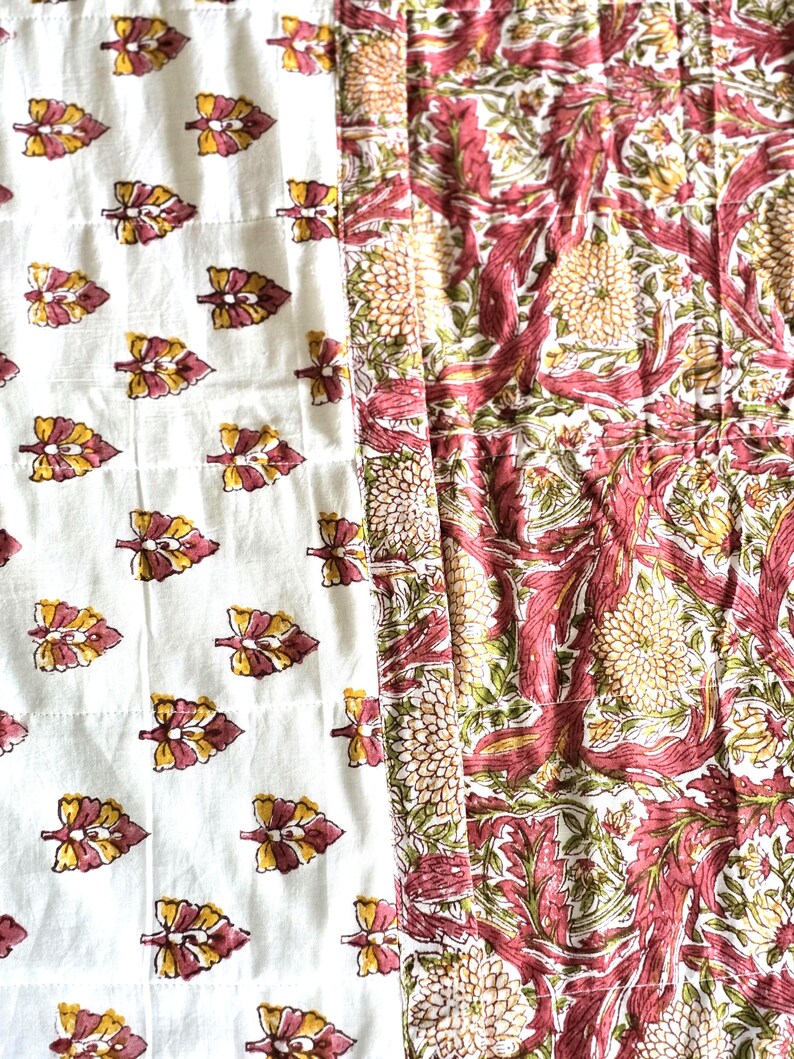 May include: A quilted textile featuring two distinct patterns. One side showcases a white background with repeating leaf motifs in yellow and maroon. The other side displays a floral design in maroon, green, and yellow.