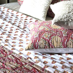 May include: A bed with a white comforter and pillows. The comforter has a white background with a repeating pattern of small yellow and red leaf shapes. The pillows are white and textured, and a patterned pillow with red and yellow floral designs.