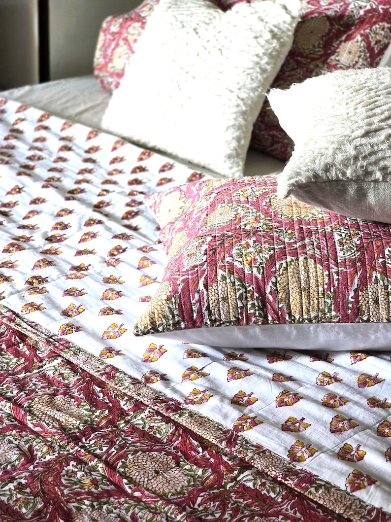 May include: A bed with a white comforter and pillows. The comforter has a white background with a repeating pattern of small yellow and red leaf shapes. The pillows are white and textured, and a patterned pillow with red and yellow floral designs.