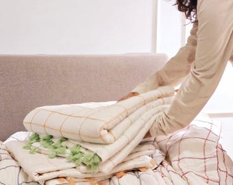 May include: A stack of folded, cream-colored quilts with grid patterns in various colors, including yellow, red, and navy blue. Some quilts have green or orange tassels. The quilts are on a light-colored sofa.