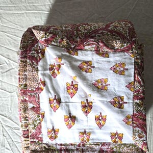May include: Folded quilt featuring a white center with a repeating pattern of pink, yellow, and orange leaves. The border and backing have a floral design in shades of pink, green, and brown.