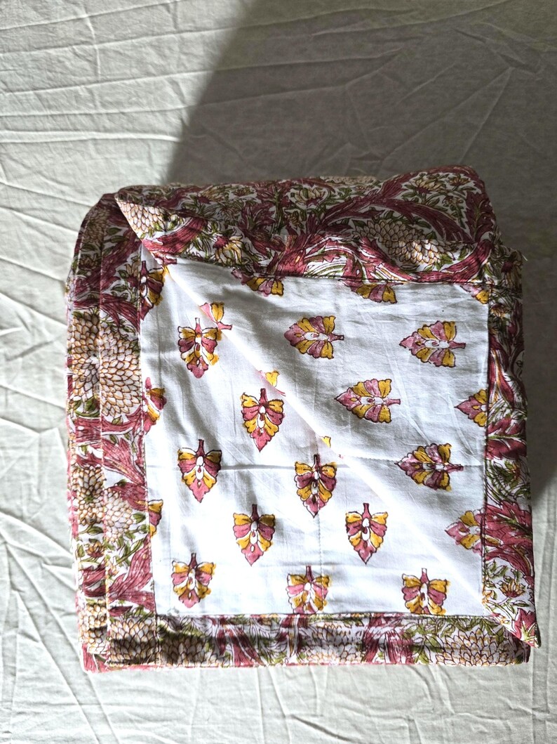 May include: Folded quilt featuring a white center with a repeating pattern of pink, yellow, and orange leaves. The border and backing have a floral design in shades of pink, green, and brown.