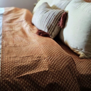 May include: A bed with a rust-coloured blanket featuring a geometric pattern. Several pillows, some patterned and some plain white, are arranged on the bed. The blanket has a repeating triangle design.