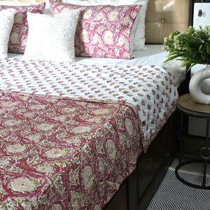 May include: A bed with a cream-colored headboard and a white bedspread. The bed is adorned with decorative pillows and a quilt featuring a floral pattern in shades of red, green, and beige. The quilt's design is repeated on the pillows.