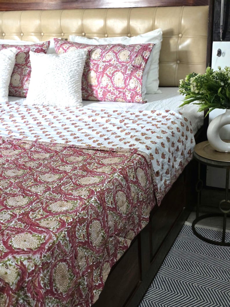 May include: A bed with a cream-colored headboard and a white bedspread. The bed is adorned with decorative pillows and a quilt featuring a floral pattern in shades of red, green, and beige. The quilt's design is repeated on the pillows.
