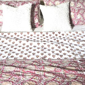 May include: A bed with white, textured pillows and patterned bedding. The bedding features a white background with small shield-like designs and a floral pattern in shades of pink and beige. The headboard is a light beige color.