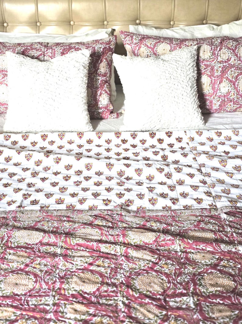 May include: A bed with white, textured pillows and patterned bedding. The bedding features a white background with small shield-like designs and a floral pattern in shades of pink and beige. The headboard is a light beige color.