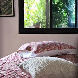 May include: A bed with a patterned red and white comforter and two pillows. One pillow has a red and white floral pattern, and the other is a fluffy white pillow. A window with green plants is in the background.