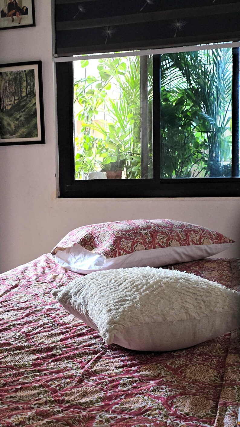 May include: A bed with a patterned red and white comforter and two pillows. One pillow has a red and white floral pattern, and the other is a fluffy white pillow. A window with green plants is in the background.