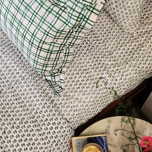 May include: Bedding with a small, dark green floral pattern. A pillow with a green plaid design rests on top. A book titled "Indigo" is on a small, round table. Pink flowers are in a vase, creating a calming aesthetic.