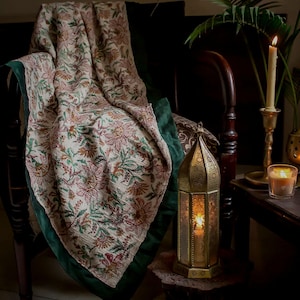 May include: A floral blanket with a green velvet trim is draped over a dark wooden chair. A gold lantern with a lit candle sits on a small table. A lit candle in a brass holder and a small plant are also visible.