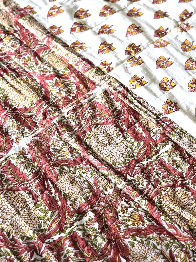 May include: A close-up of a white and red floral patterned quilt. The quilt features a repeating pattern of large, stylized flowers and leaves in shades of red, pink, and green. The top section has a white background with a repeating pattern of yellow and pink leaves.