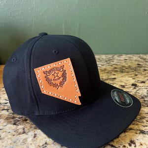 May include: Black fitted baseball cap with a brown leather patch featuring the state of Nevada outline and the state seal. The patch is stitched onto the cap.