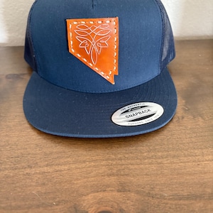 May include: Navy blue trucker hat with a curved brim and mesh back. Features a leather patch in the shape of Nevada, with a white stitched design. The hat has a snapback closure and a "Snapback" tag.