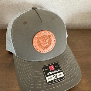 May include: A gray Richardson 112 trucker hat with a white mesh back and a brown leather patch. The patch features a star and text. The hat is adjustable.
