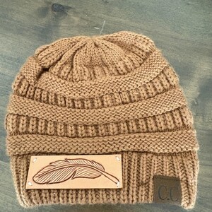 May include: A tan knit beanie with a leather patch featuring a feather design. The hat has a ribbed knit texture and a small, square tag with the letters "C.C." The hat is a warm, casual accessory.