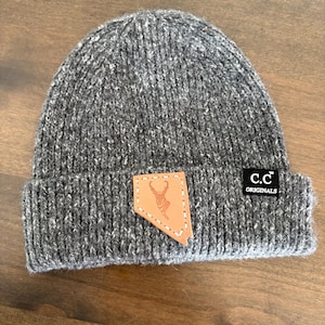 May include: A gray knit beanie with a leather patch featuring a deer silhouette within a state outline. The beanie has a folded brim and a small black tag that reads "c.c ORIGINALS". The hat is made of a soft, textured material.