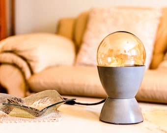 May include: A gray table lamp with a clear glass globe shade and a warm light bulb. The lamp is on a white table with a decorative glass bowl.