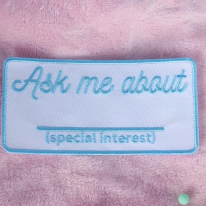 Puede incluir: Un parche blanco con texto bordado en azul que dice "Ask me about (special interest)".