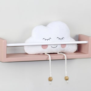 Puede incluir: Una estantería de madera rosa con una barra blanca, que sostiene un peluche blanco con forma de nube. La nube tiene ojos bordados y una boca sonriente, con piernas de cuerda colgantes y cuentas de madera. Un artículo decorativo para una guardería.