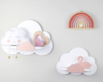 May include: White cloud-shaped shelves with pink accents and a rainbow wall hanging. One shelf features a plush cloud with a smiling face, another has a heart, and the third has a swan. The rainbow is red, pink, and gold.