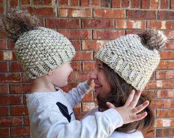 May include: Two people wearing matching knit hats with faux fur pom poms. The hats are a light beige color with a speckled pattern.