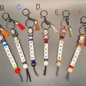 May include: Keychains with horror movie character charms and beaded accents. Each keychain features a different character, including Chucky, Ghostface, Jason Voorhees, Freddy Krueger, and Pennywise. The keychains have silver-tone hardware and colorful tassels.