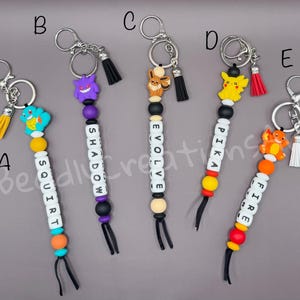 May include: Five keychains labeled A through E, each featuring a silicone character charm, colorful round beads, and white letter beads spelling SQUIRT, SHADOW, EVOLVE, PIKA, and FIRE. Each keychain includes a metal ring and a small colored tassel.