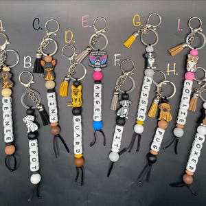 May include: A collection of dog-themed keychains. Each keychain features a dog charm, beads, and a tassel. The keychains are labeled with dog names like "Weenie", "Spot", "Tiny", "Guard", "Angel", "Pit", "Aussie", "Pug", "Collie", and "Boxer".