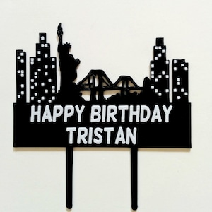 May include: Black and white cake topper featuring a New York City skyline with the Statue of Liberty, a bridge, and skyscrapers. The topper also includes the text "Happy Birthday Tristan".