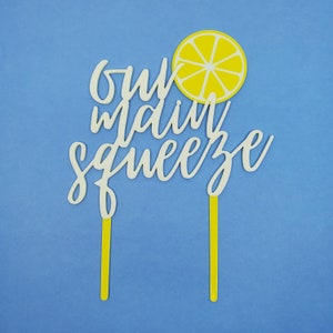 May include: White cake topper with the text "our main squeeze" and a yellow lemon slice.