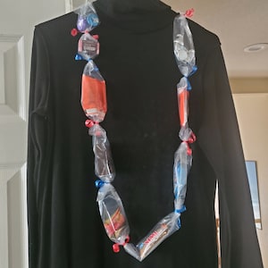 May include: A black mock neck shirt with a colorful candy lei made of plastic bags tied together with red and blue ribbon. The candy is wrapped in clear plastic and includes chocolate, candy bars, and other treats.