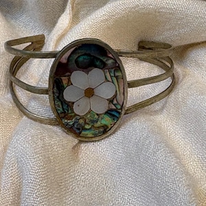 May include: A cuff bracelet featuring a triple-band metal frame and a central oval inlay. The inlay displays a white six-petaled flower with a gold centre, set against a background of iridescent, multi-coloured abalone shell fragments.