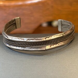 May include: A silver-toned metal cuff bracelet featuring a central twisted rope texture flanked by two smooth bands with stamped wavy line patterns. The bracelet has an open back design with flat, squared-off ends and a polished metallic finish.