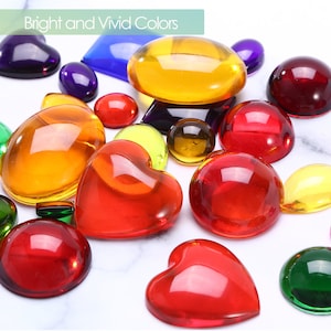 May include: Assortment of colorful, clear glass cabochons in various shapes, including hearts, circles, and ovals. The cabochons are arranged on a white surface.