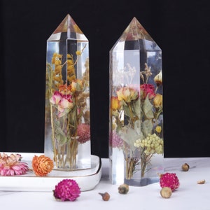 May include: Two clear crystal prisms with dried flowers and greenery inside. The prisms are on a white surface with dried flowers scattered around them.