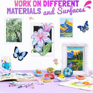 May include: An assortment of painted rocks, framed artwork, and a decorated glass are displayed on a purple surface. The artwork includes a landscape, a flower, and a tree scene. The text at the top reads "WORK ON DIFFERENT MATERIALS and Surfaces."