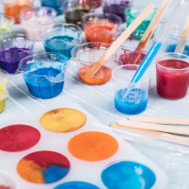 May include: A white plastic mold with eight round cavities for making resin crafts. The cavities are filled with colorful resin, including blue, orange, yellow, and red. There are also several small cups filled with different colored resin, including blue, red, orange, and yellow.