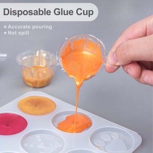 May include: A clear plastic disposable glue cup with orange glue being poured into a silicone mold with six circular depressions. The mold has a paw print design in one of the depressions. The text "Disposable Glue Cup" is at the top of the image. The text "Accurate pouring" and "Not spill" are listed below the title.