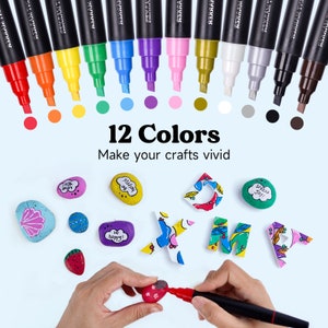 May include: Set of 12 paint markers with black caps and colorful paint tips. The markers are arranged in a row with the tips facing the viewer. The colors are red, orange, yellow, green, blue, purple, pink, gold, white, gray, black, and brown. The text "12 Colors" and "Make your crafts vivid" is displayed below the markers. The image also shows painted rocks and letters.