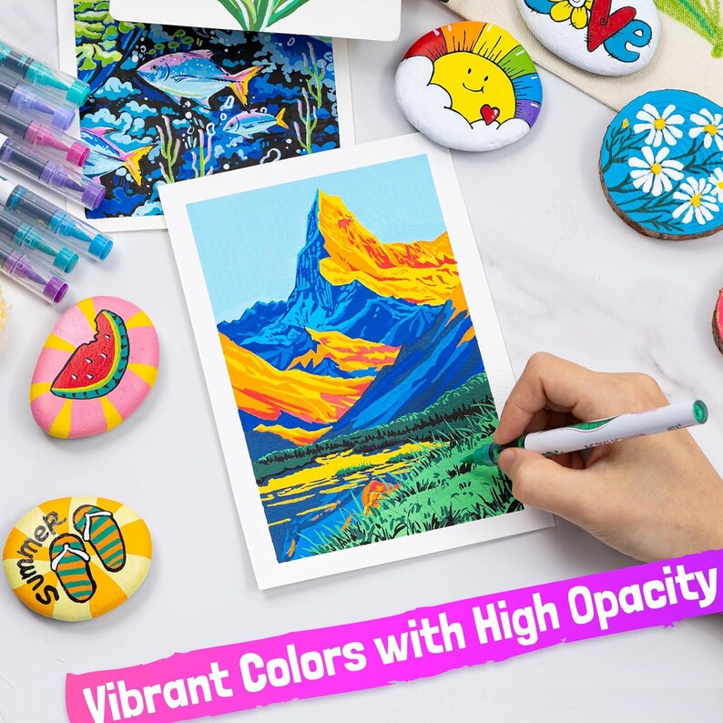 May include: A vibrant image featuring art supplies and painted rocks. A hand uses a green marker to color a landscape painting with blue, yellow, and green hues. The text "Vibrant Colors with High Opacity" is displayed. Other painted rocks feature summer-themed designs.