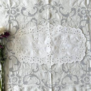 May include: A white, oval-shaped, eyelet lace doily with floral cutouts rests on a white, patterned fabric. A small bouquet of purple and white flowers is positioned to the left of the doily, adding a touch of color.