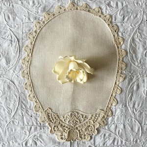 May include: An oval-shaped beige linen doily featuring an intricate cream-colored lace border with scalloped edges. Centered on the doily is a single, realistic cream-colored daffodil flower with a yellow center, resting flat against the fabric.