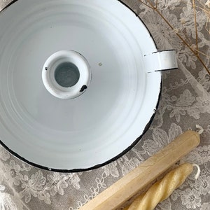 May include: White enamel candle holder with a black rim and a single candle holder in the center. The holder is sitting on a white lace tablecloth with two candles next to it.