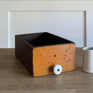 May include: A wooden box with a dark brown exterior and a bright orange interior. The box has a white knob on the front and is sitting on a wooden surface. A white ceramic pitcher is next to the box.